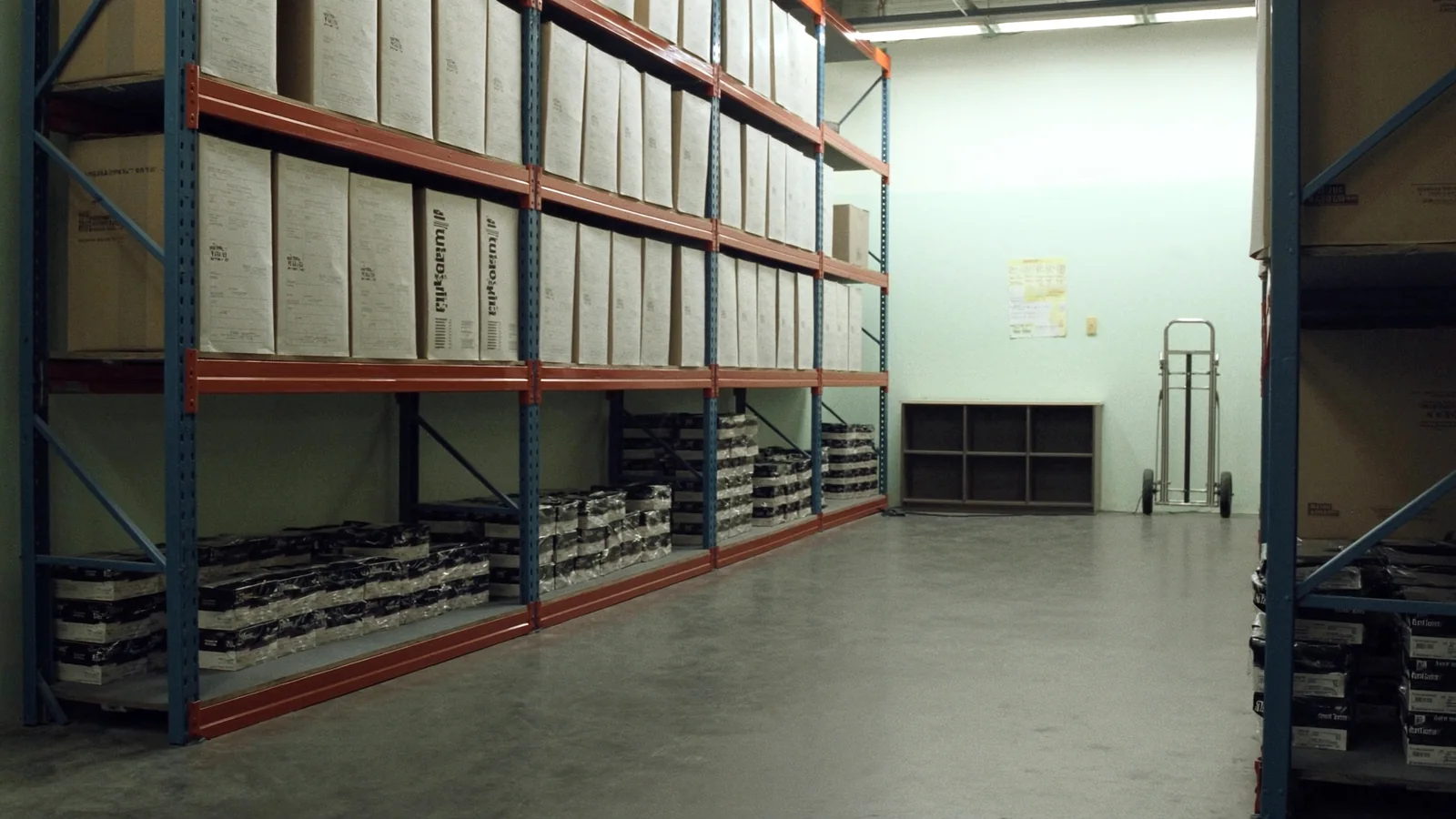An aisle of steel pallet racking in a small office supply warehouse stacked with cartons of copier paper, a hand truck parked at the end of the run.