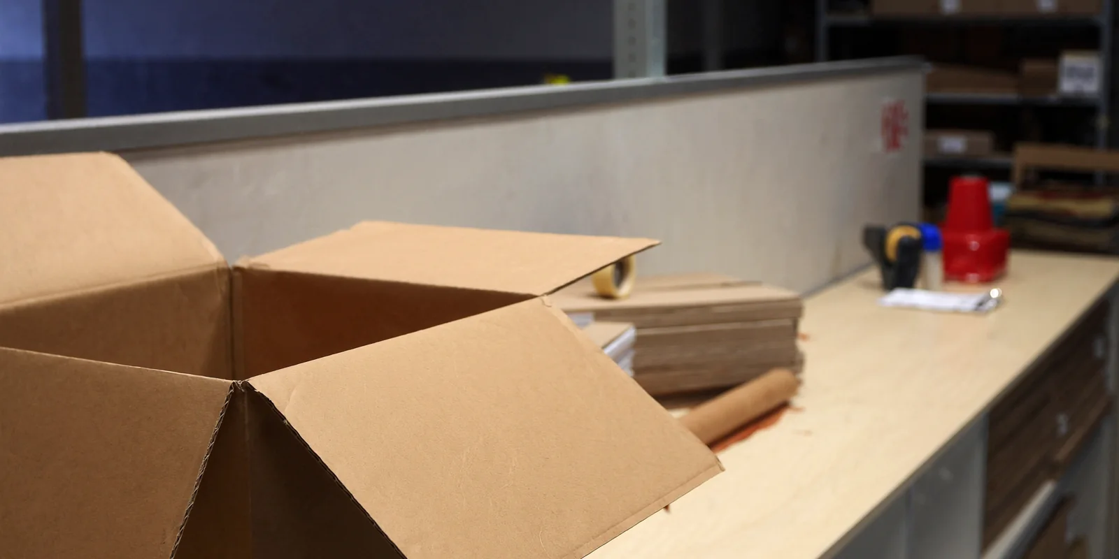 An order picking bench in a warehouse with a part filled carton, a clipboard, a tape gun and a stack of flat boxes, racking blurred behind.