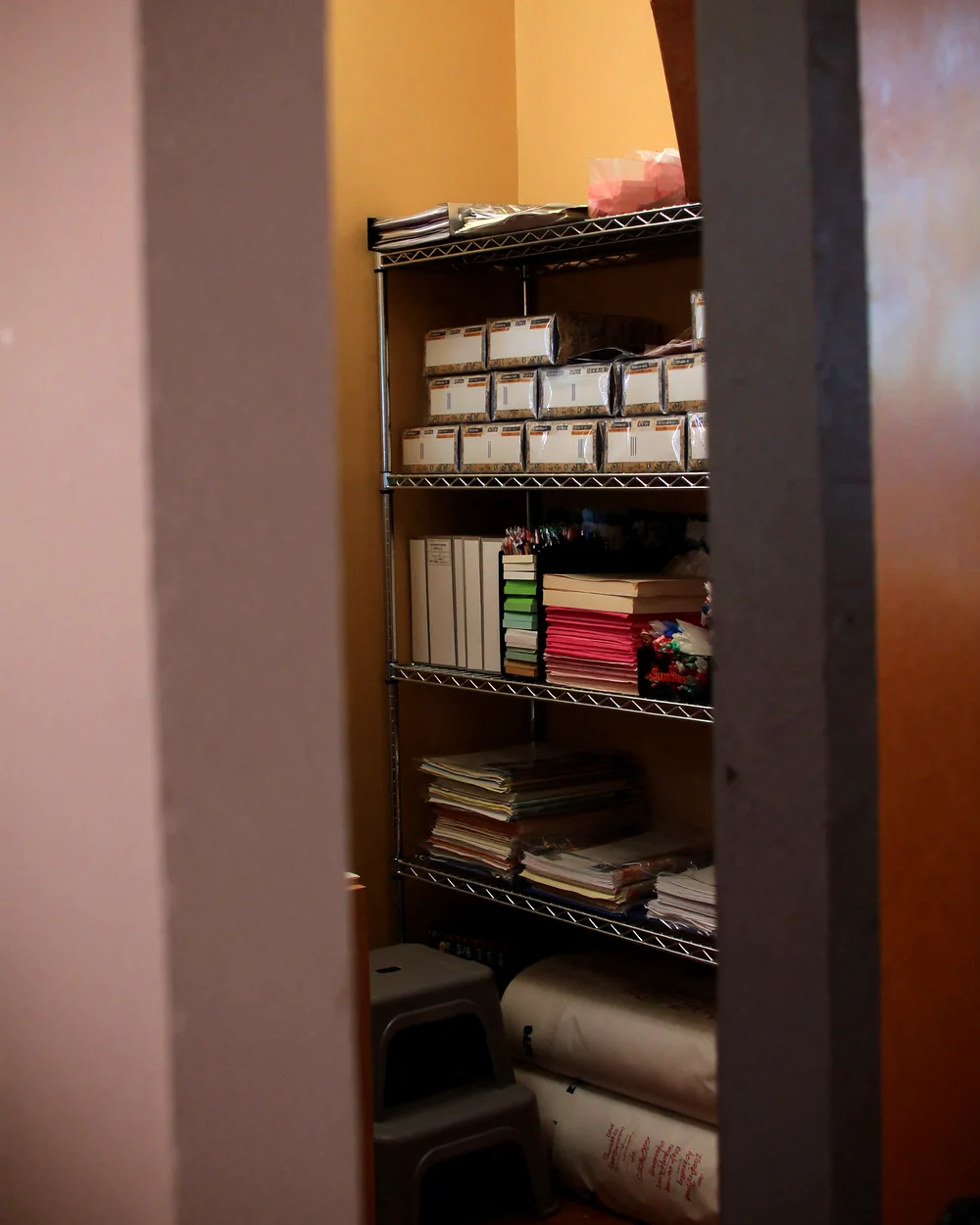 An open office supply closet with wire shelving holding boxed pens, pads, folders and reams of paper, squared up and facing forward.
