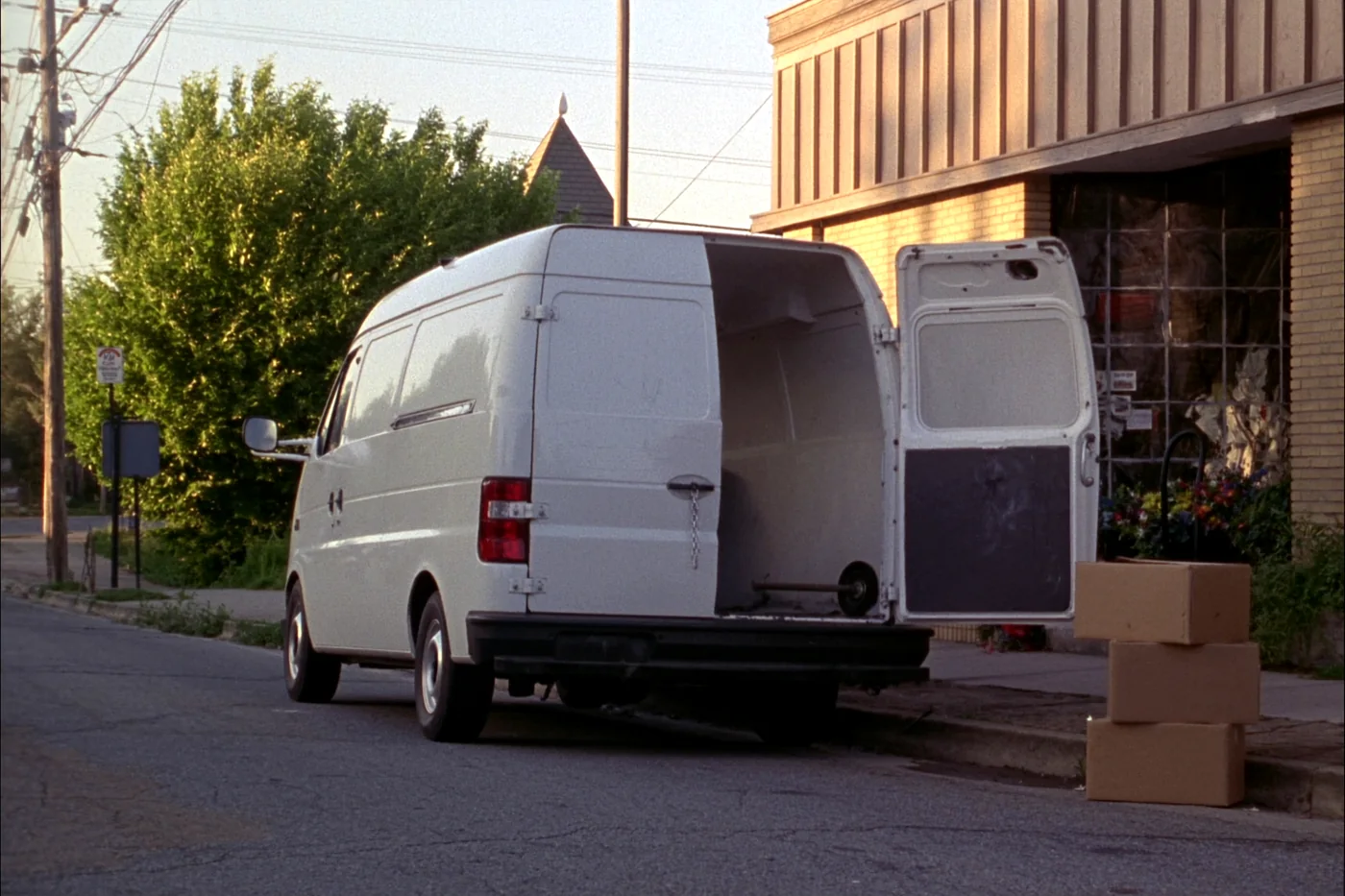 A plain white cargo van at the kerb outside a small brick office building with its side door open, three cartons and a hand truck on the pavement.
